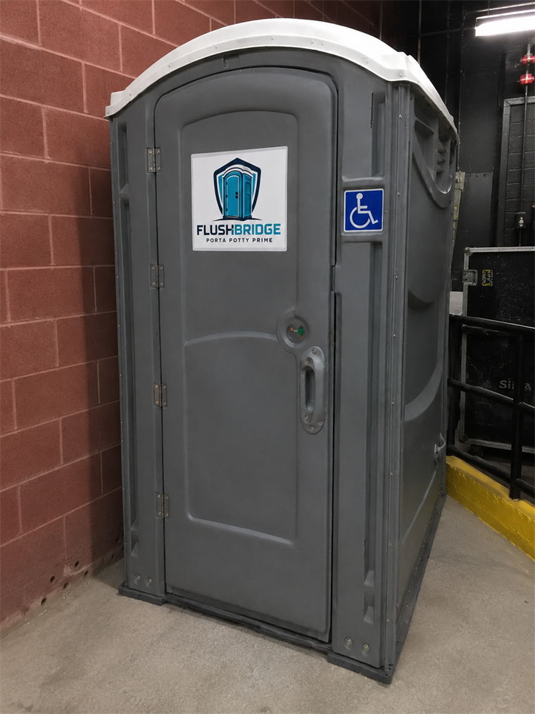 Gray ADA accessible portable restroom with a wheelchair decal, standing against a brick wall
