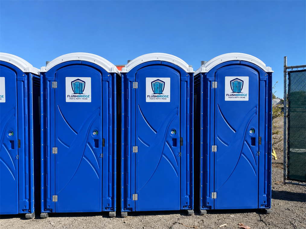 Four blue portable toilets lined up in a row on gravel beside a fence