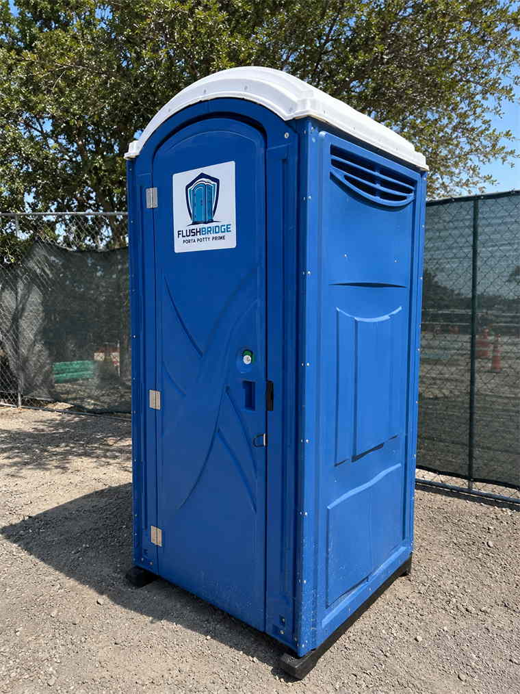 Blue Flushbridge Porta Potty Prime portable toilet standing on gravel beside a chain-link fence