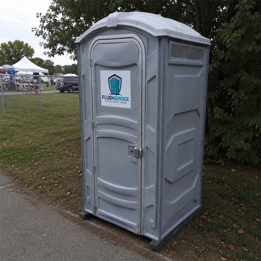 Gray portable toilet standing on grass, with event tents in the background