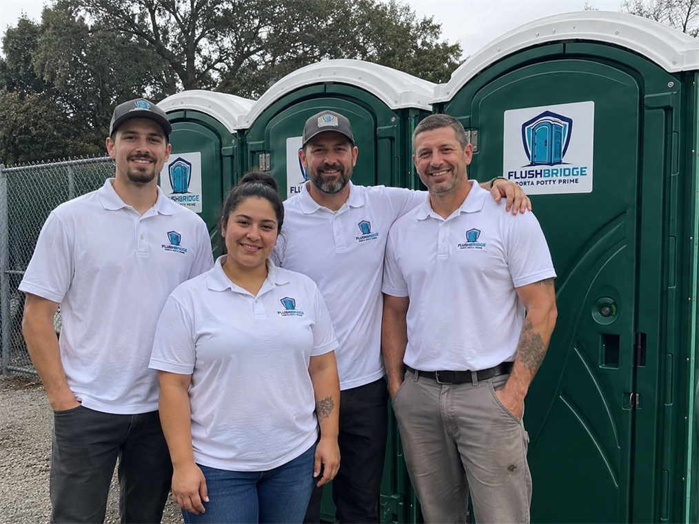 Four Flushbridge Porta Potty Prime team members in white polo shirts standing in front of portable toilets
