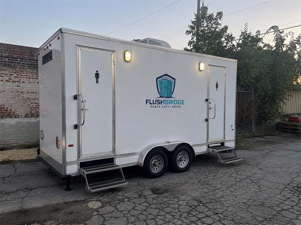 Side view of a white restroom trailer parked on asphalt beside a brick wall